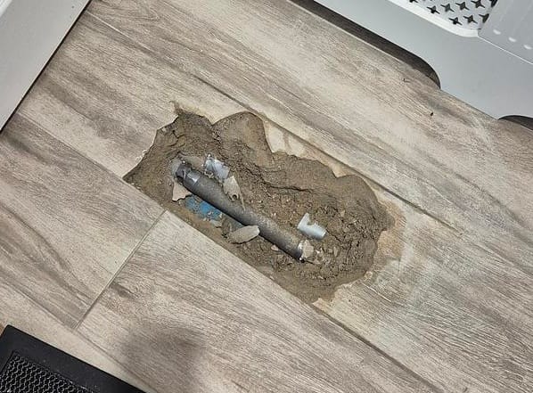 Small opening in wood-effect floor tiles exposing pipework