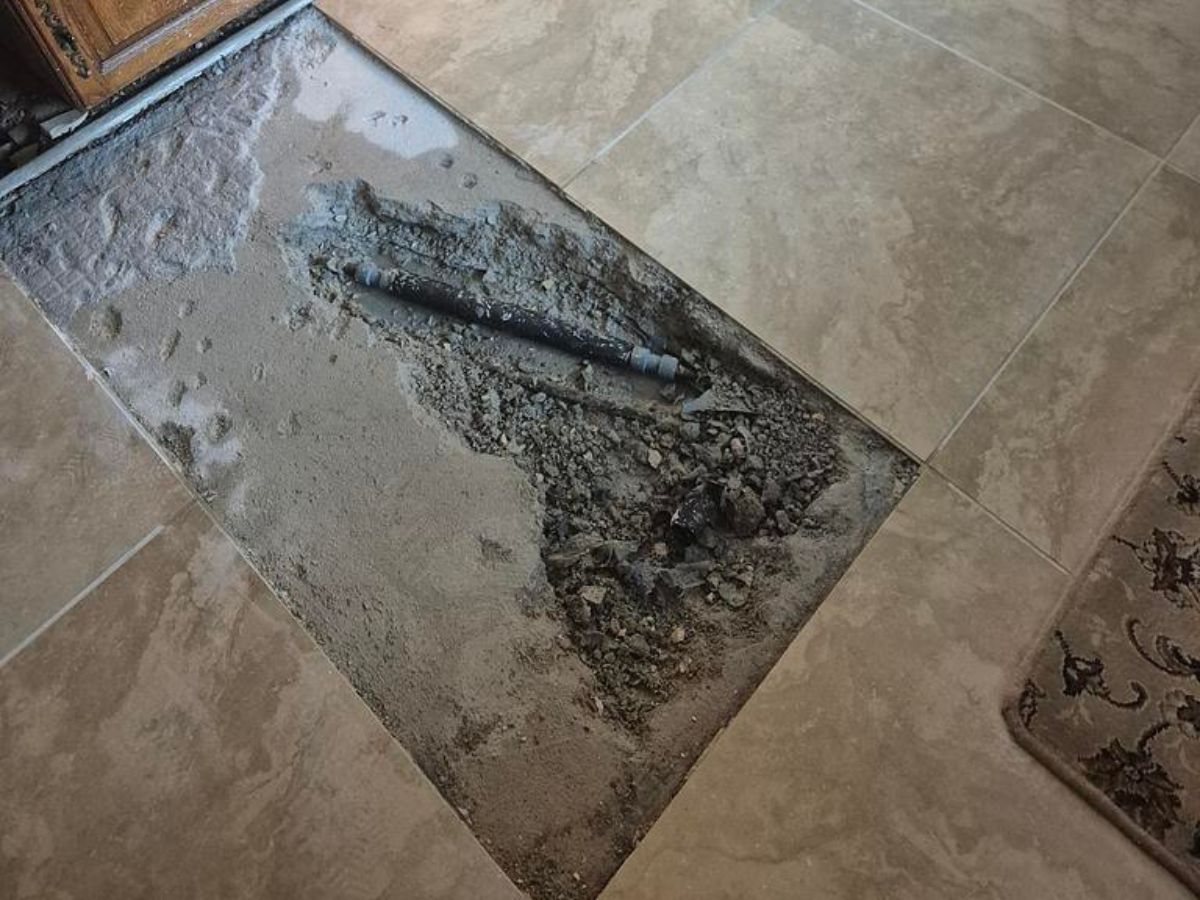 Pipe exposed through a rectangular opening in beige floor tiles
